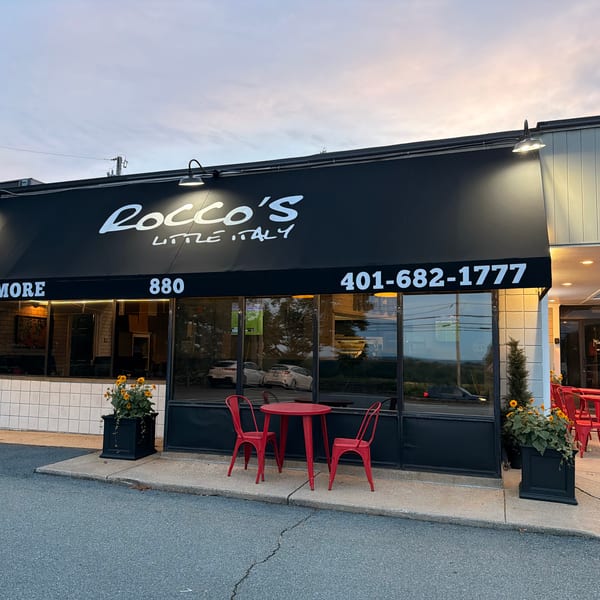 Exterior of Rocco's Little Italy with a black awning, red outdoor seating, and flower planters at dusk.