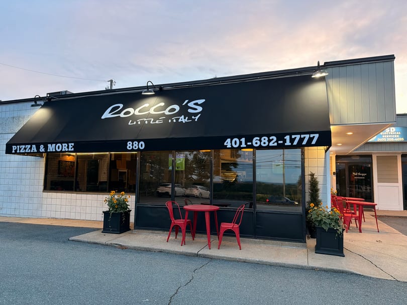 Exterior of Rocco's Little Italy with a black awning, red outdoor seating, and flower planters at dusk.