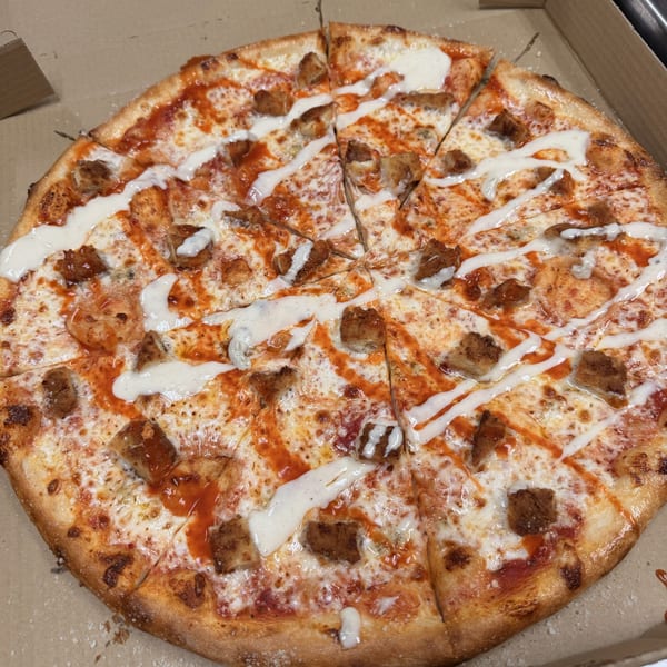 A buffalo chicken pizza with white and orange sauce drizzled over it, in a cardboard box.