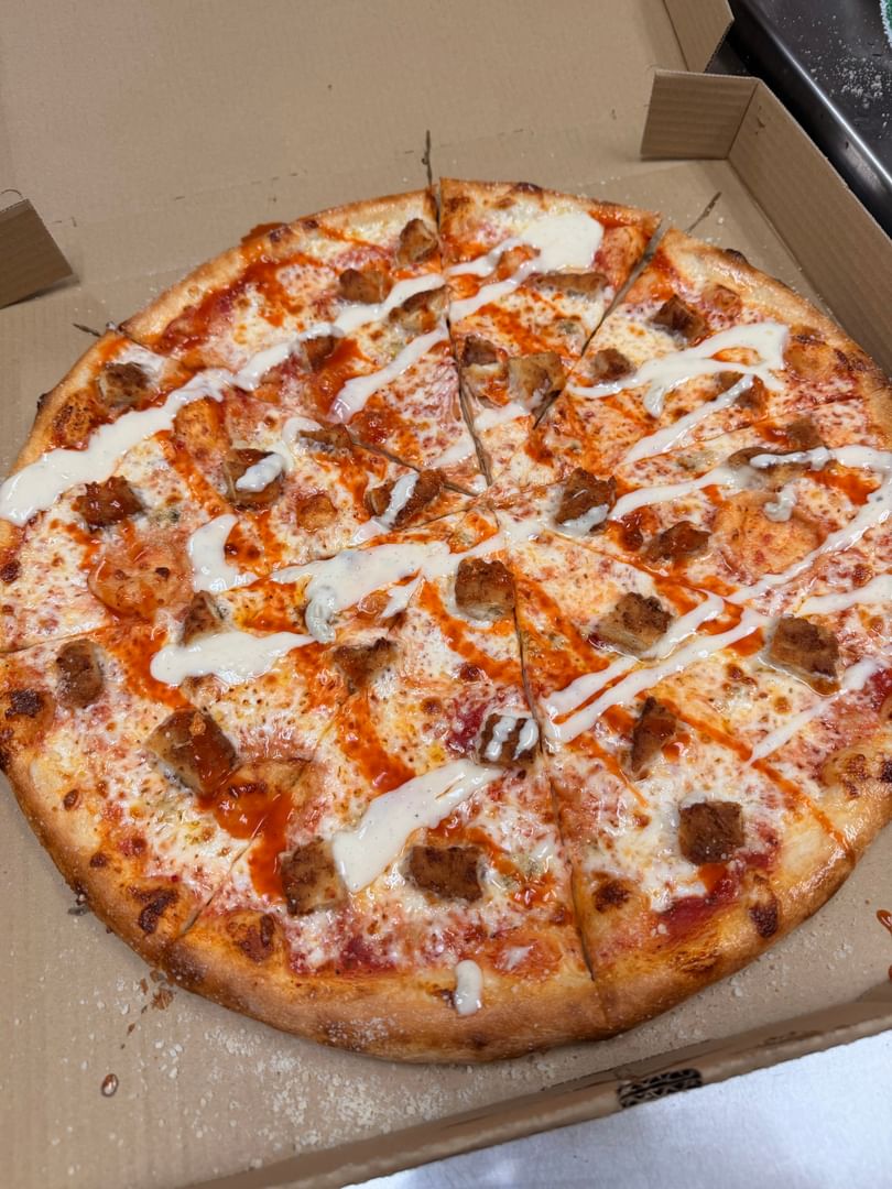 A buffalo chicken pizza with white and orange sauce drizzled over it, in a cardboard box.