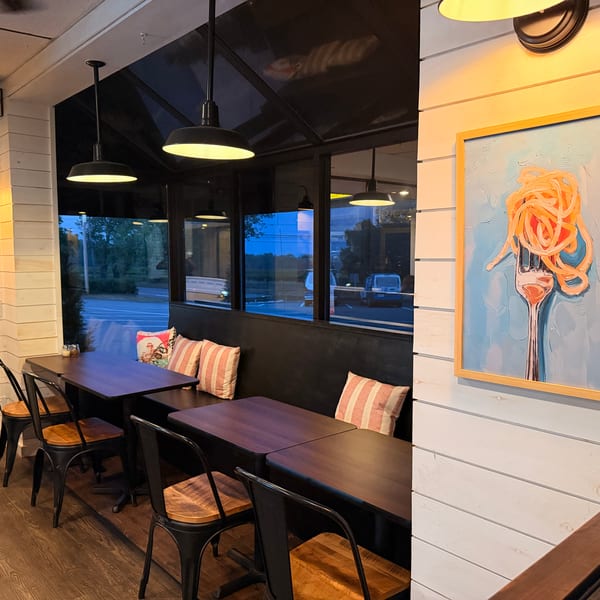 Cozy restaurant interior with dark tables, metal chairs, striped pillows, and pasta-themed artwork on light walls.