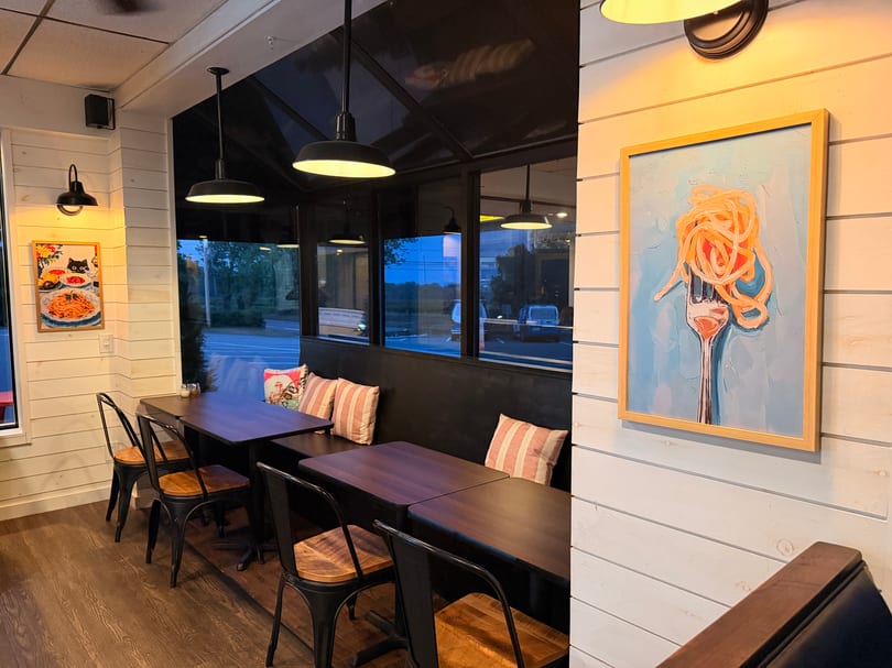 Cozy restaurant interior with dark tables, metal chairs, striped pillows, and pasta-themed artwork on light walls.