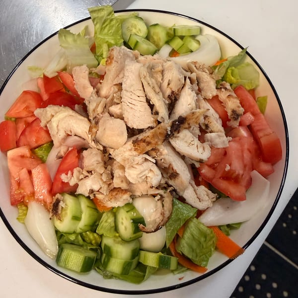 A large salad with grilled chicken, lettuce, tomatoes, cucumbers, and onions in a white bowl.