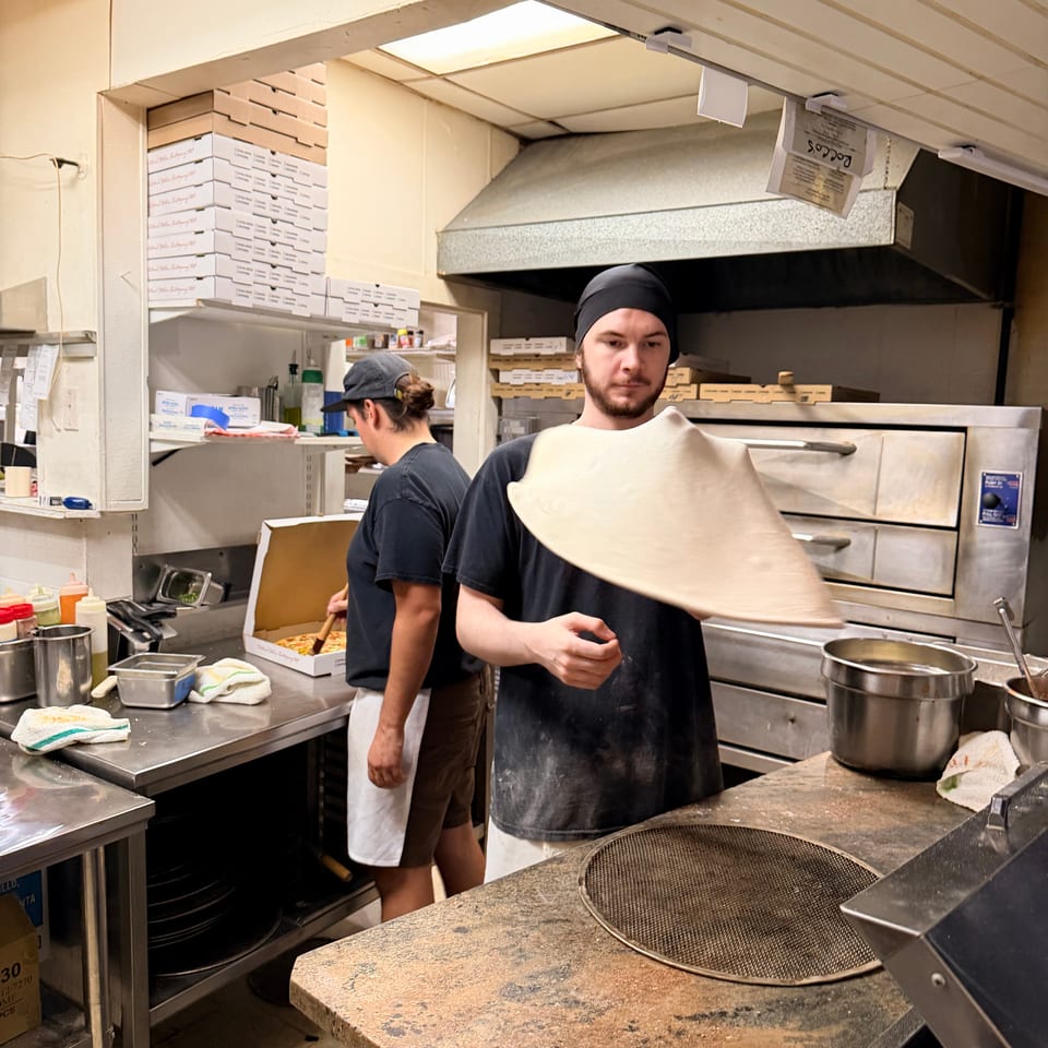 Two restaurant workers prepare food in a busy commercial kitchen surrounded by pizza boxes, stainless-steel equipment, and cooking supplies.